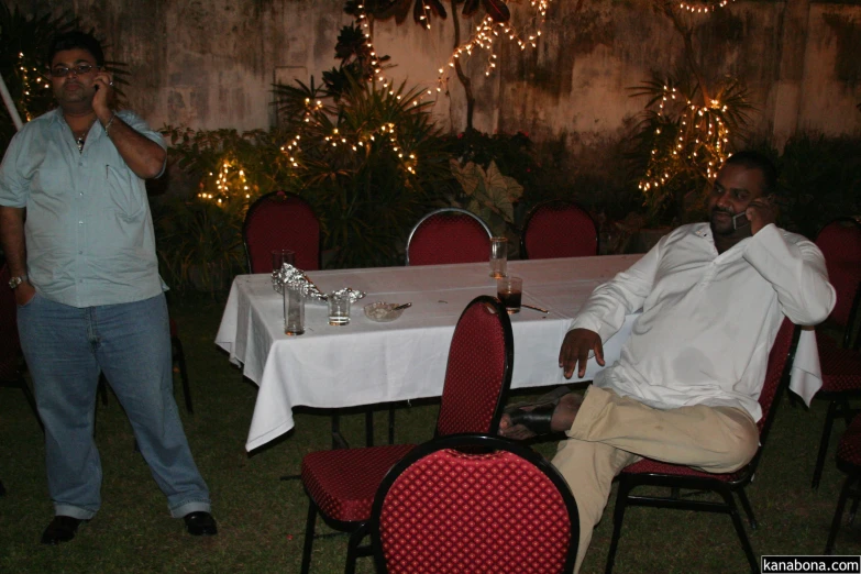 two men standing and sitting around a table outside