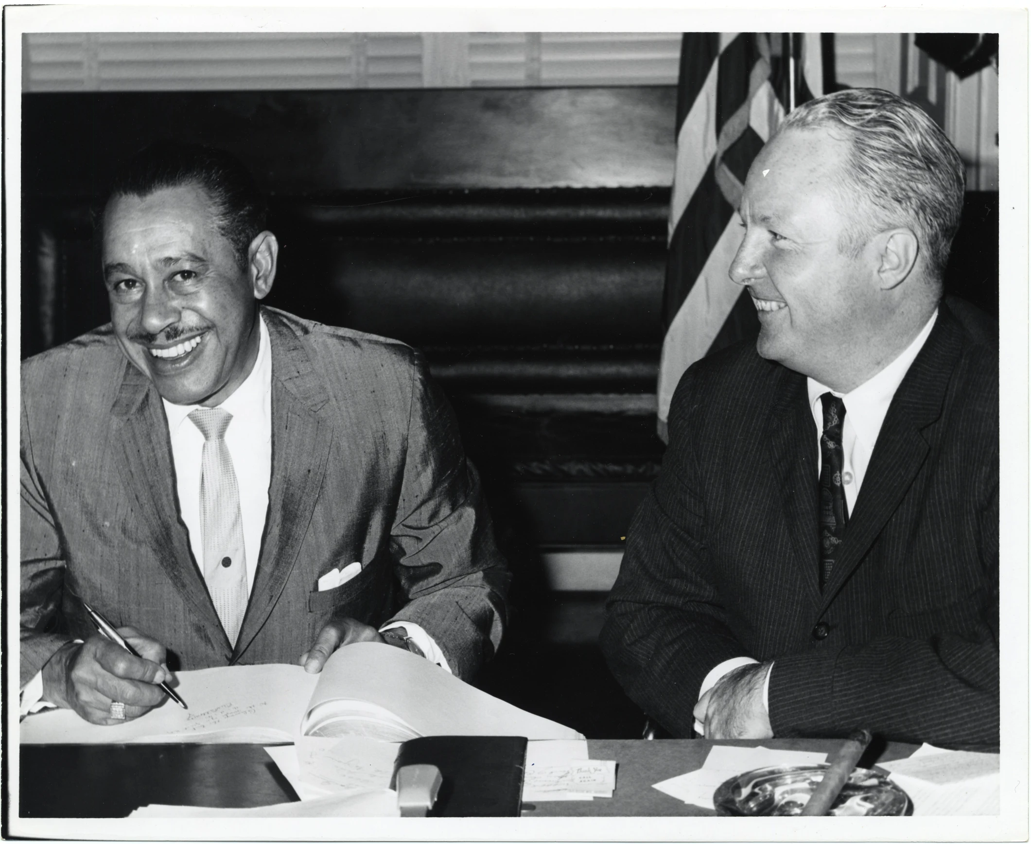 two men are sitting at a table with paper and pens