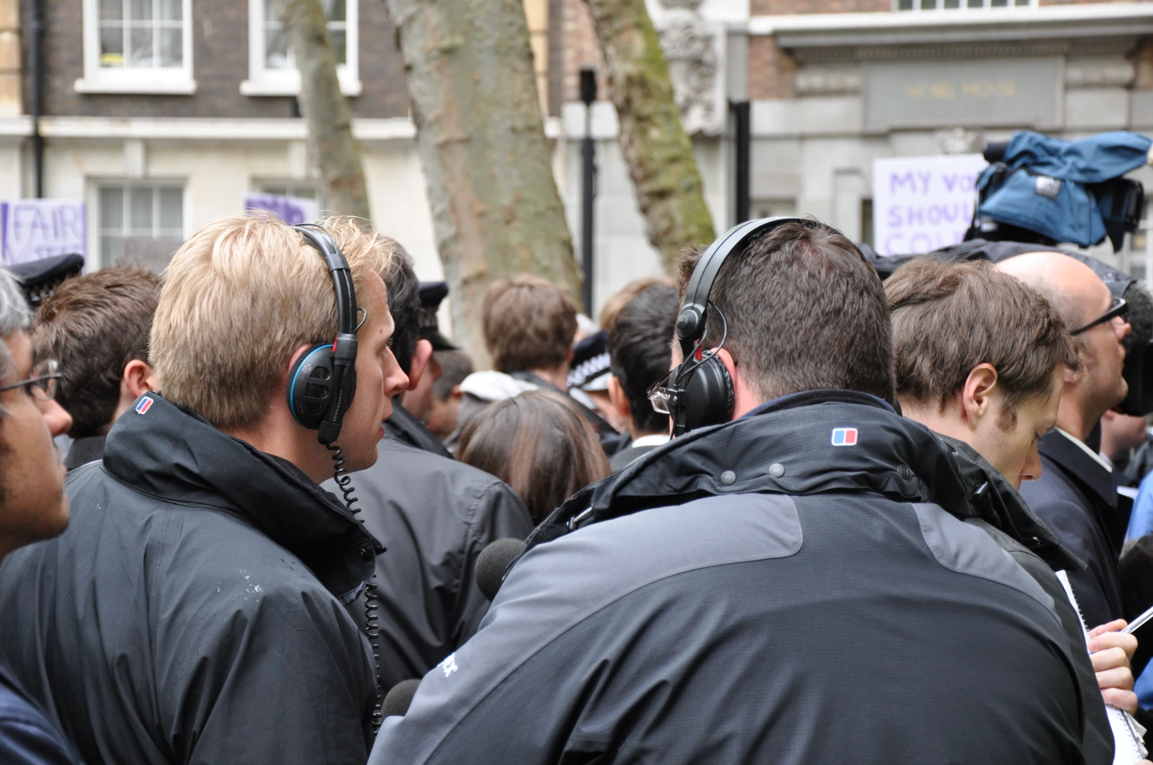 man in black jacket and headphones speaking with a crowd behind him