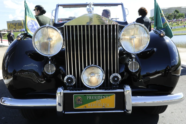 a classic car sits parked while several people walk by