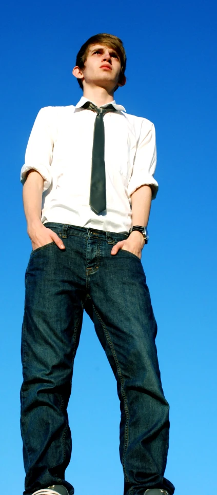 a young man standing against a blue sky with his hands in his pockets