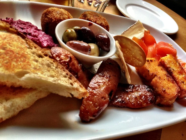 the plate has sausage, bread, tomato, olives and a knife