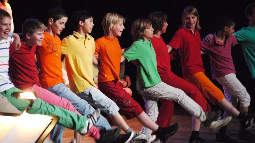 young s with green and red tops stand around while they watch as another boy holds a guitar and sings