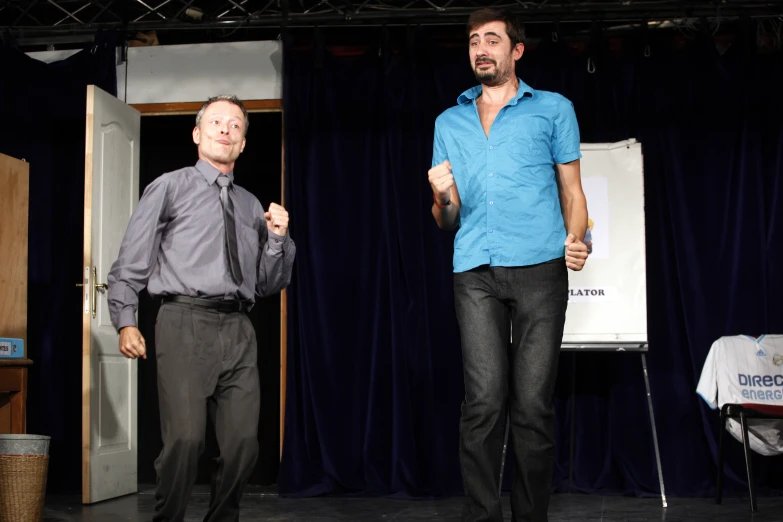 two men stand on a stage one in a tie and the other in a pants