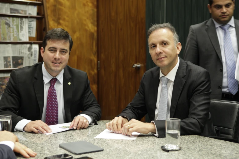 two men wearing suits sit at a table with papers