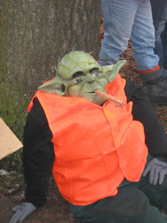 a costumed figure sitting by a tree wearing an orange vest