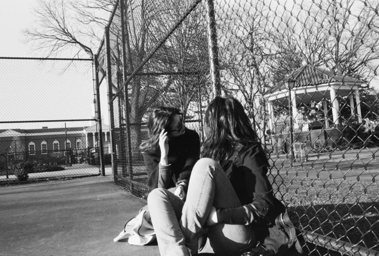 two women sit against the fence near a baseball field