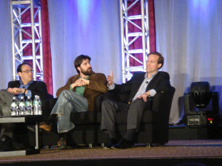 two men sitting on a stage talking to other men