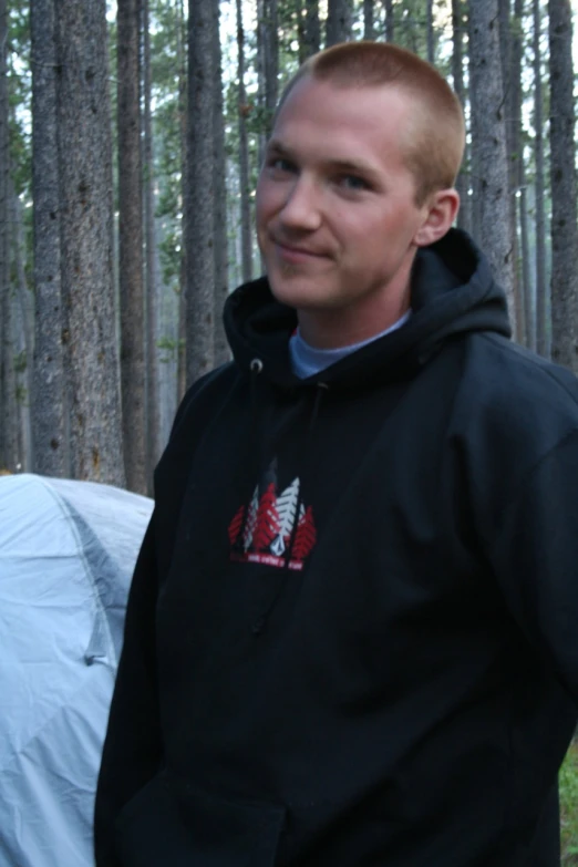 a man in a black sweatshirt stands in front of trees and is wearing his name shirt