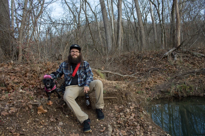 a bearded man sits with a dog by a creek