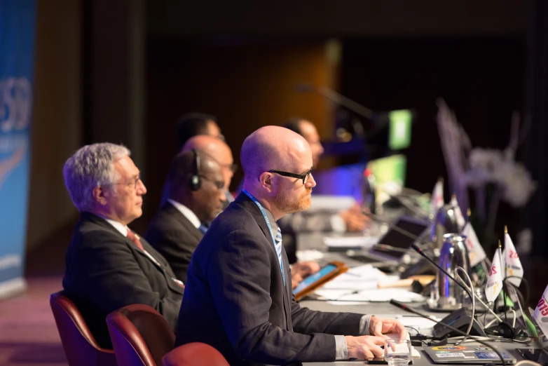 people in business suits are seated at a table with laptops and papers