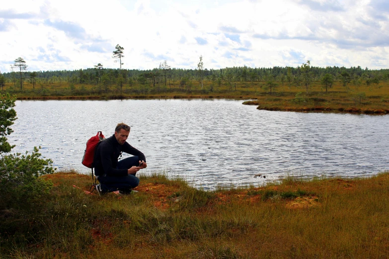 a man with a red backpack squatting by the edge of a lake