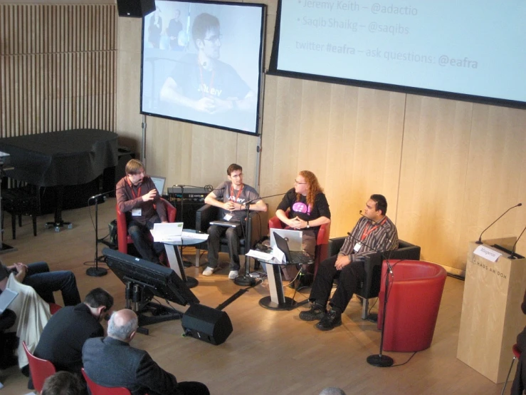 three people sitting in red chairs at the front of a room and talking