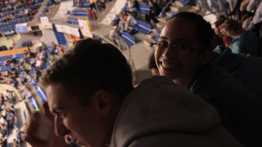 two people sitting at a basketball court with their hands up and a man looking up