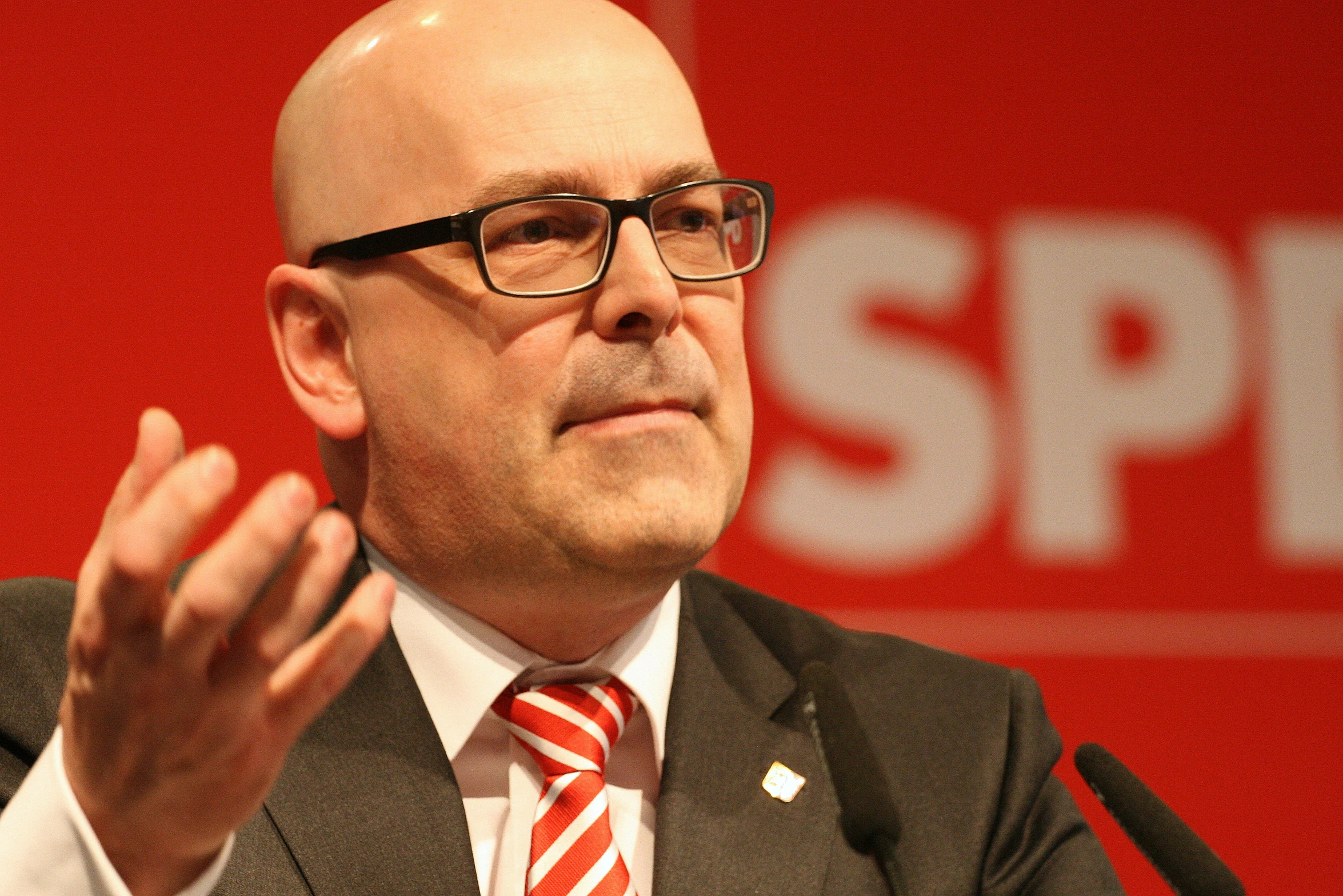 a bald man in glasses and a tie sitting in front of a red wall