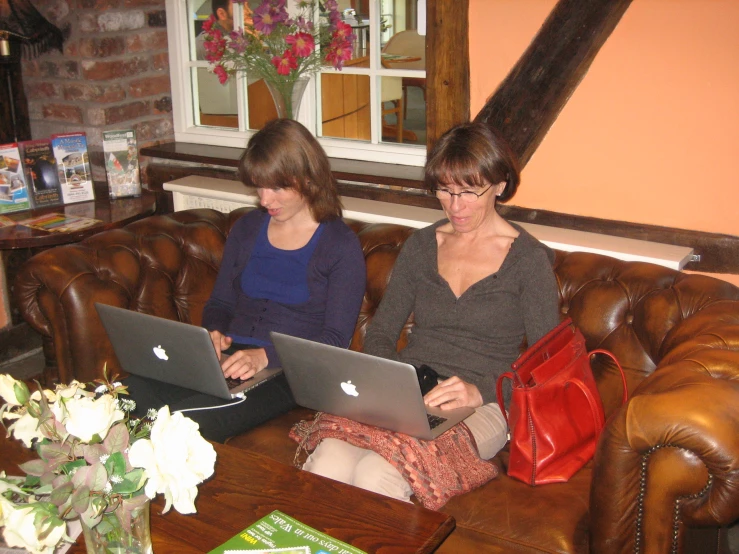 two women sit on a brown couch with their laptops