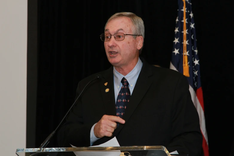 man standing at a podium wearing glasses speaking