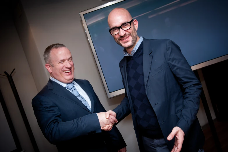 two men in suits shake hands in front of a television