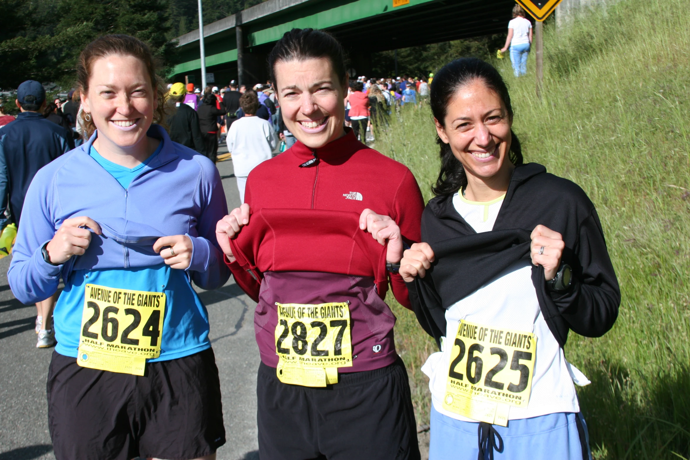 three women smiling at the camera, while standing near a road