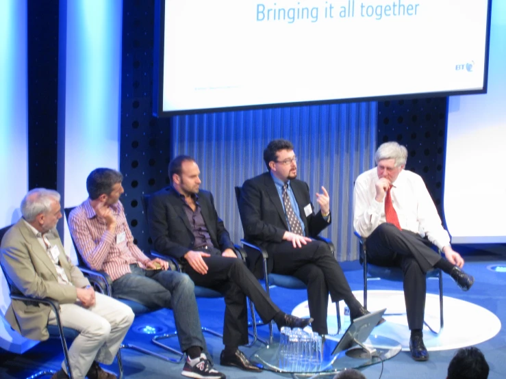 five business men sitting on stage with one man standing up