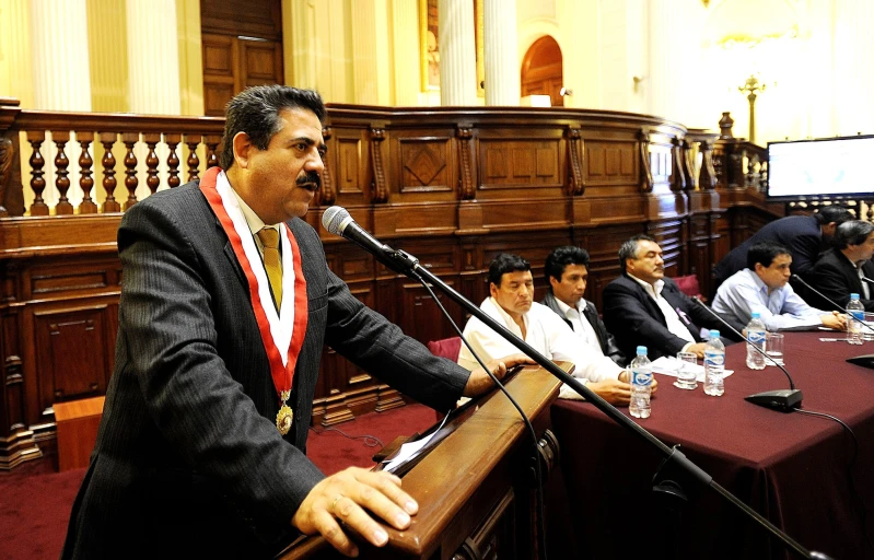 several men in suits sitting at long tables, one speaking on a microphone