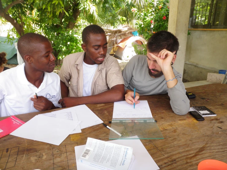 three men sit at a table and look up at papers on the table