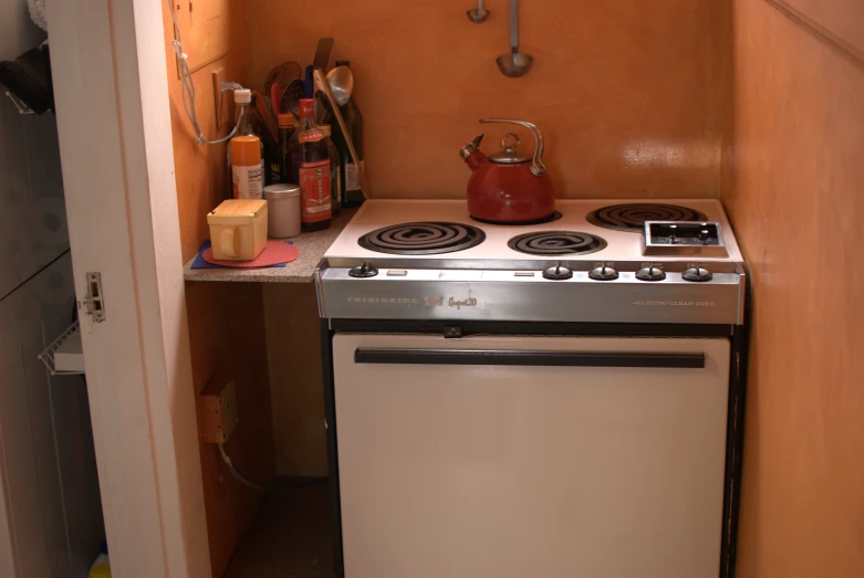 an open small kitchen with oven, tea kettle, bottles on counter and cupboards