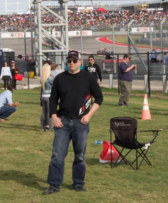 a man with a hat, sunglasses and black shirt standing in the grass near other people