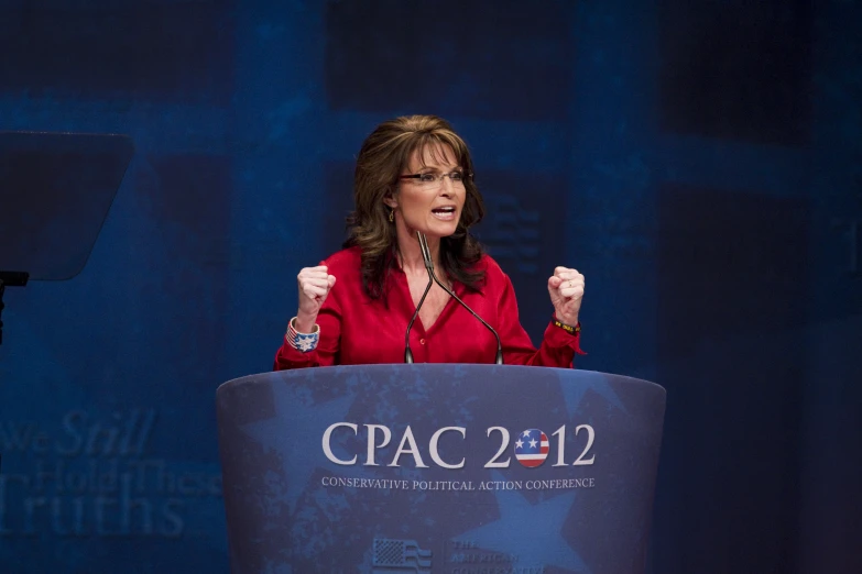 a woman speaking in front of a podium with two hands up