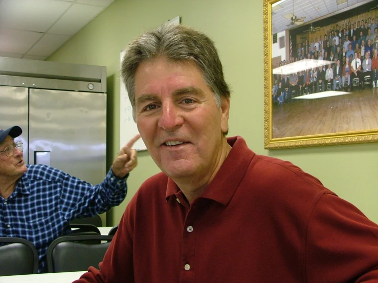 two people, one standing at the desk, one sitting behind a refrigerator