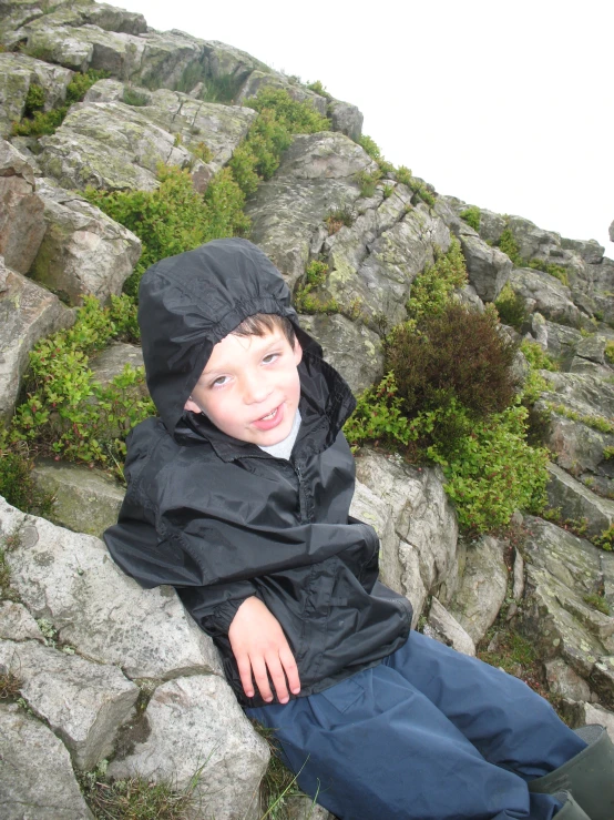 boy lying on a rocky mountain side wearing a black rain coat