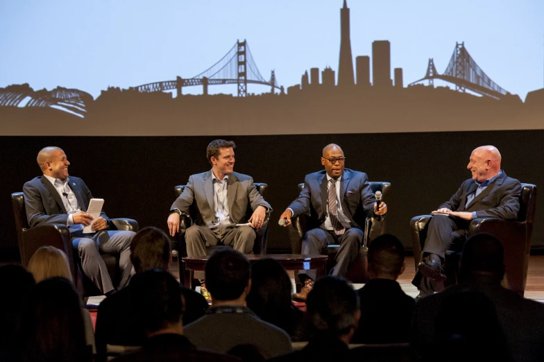 the four men sit together in chairs and look toward a large screen