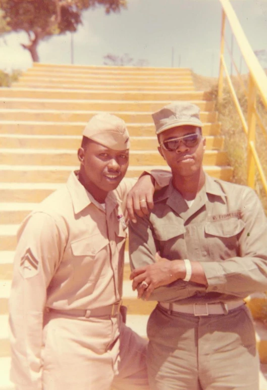 two men wearing fatigues posing in front of a set of stairs