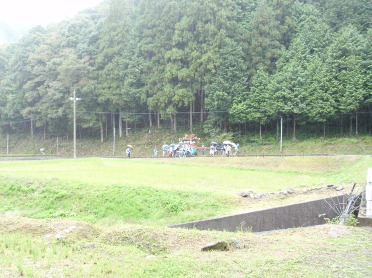 people standing outside of a green fence in front of a large tree covered hill