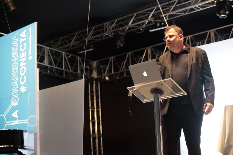 a man with glasses stands at a podium and speaks on his laptop