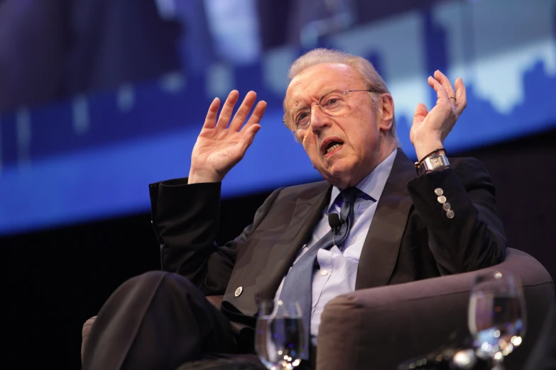 man in suit and tie sitting with both hands extended, gesturing to someone