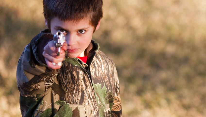 a boy in camouflage holding a gun at the camera