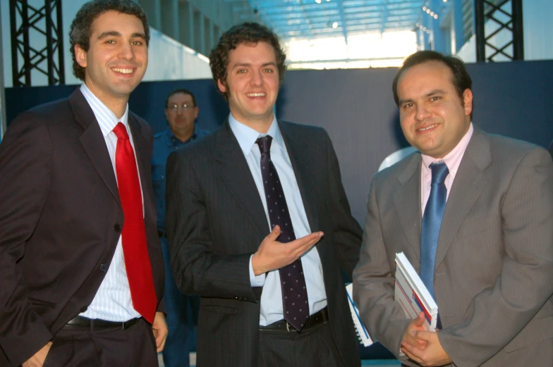 three men in business attire stand with one holding folder and another smiling at the camera