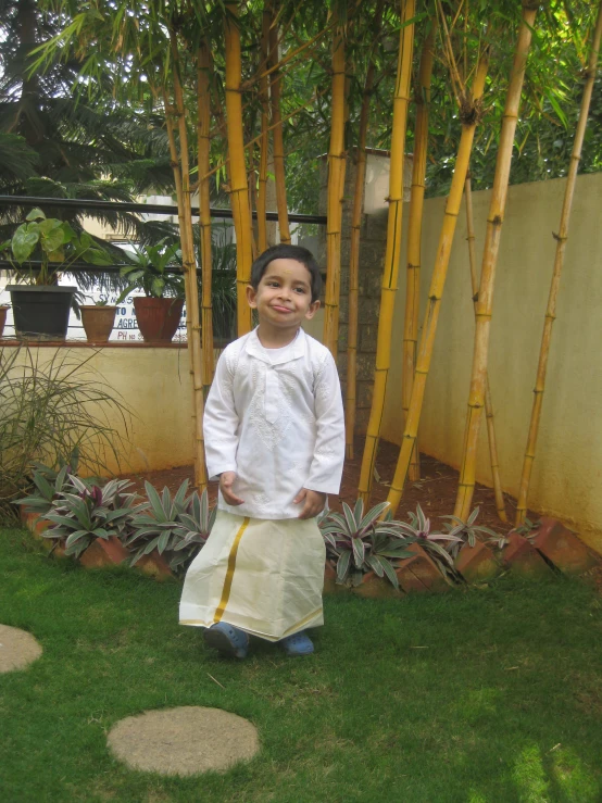 a little boy dressed in a thai dress standing on grass near a tree