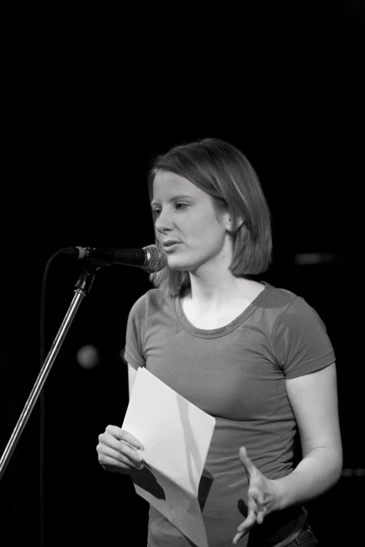 a woman in grey shirt standing at microphone with papers