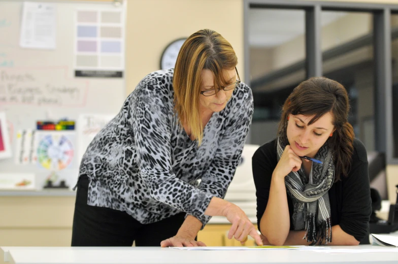 two women in the classroom one writing and one looking at the book on the table