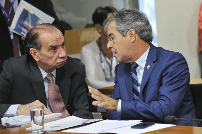 two men sitting at a table and one is on his cell phone