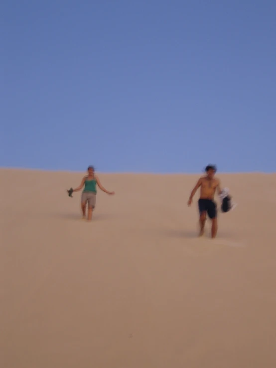 two people walking on sand dunes, the man in green is holding a surfboard