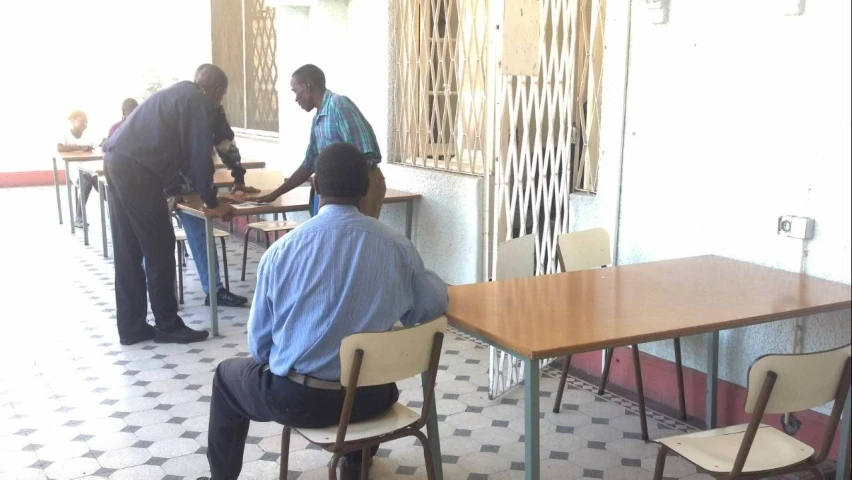 three men stand and sit in front of wooden tables