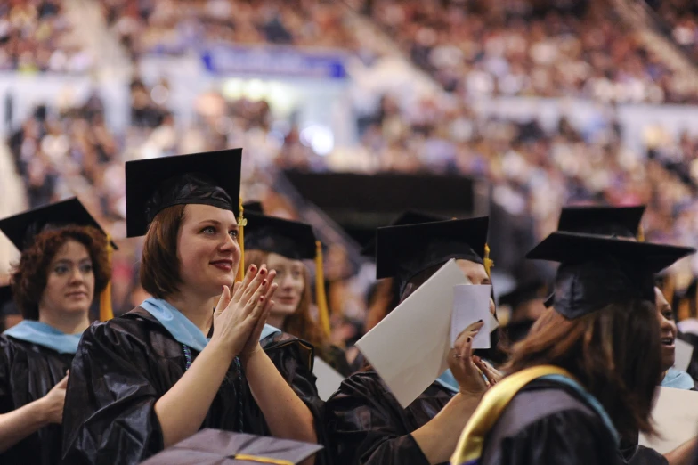 group of students from a college wearing graduation gowns clapping at graduation ceremony