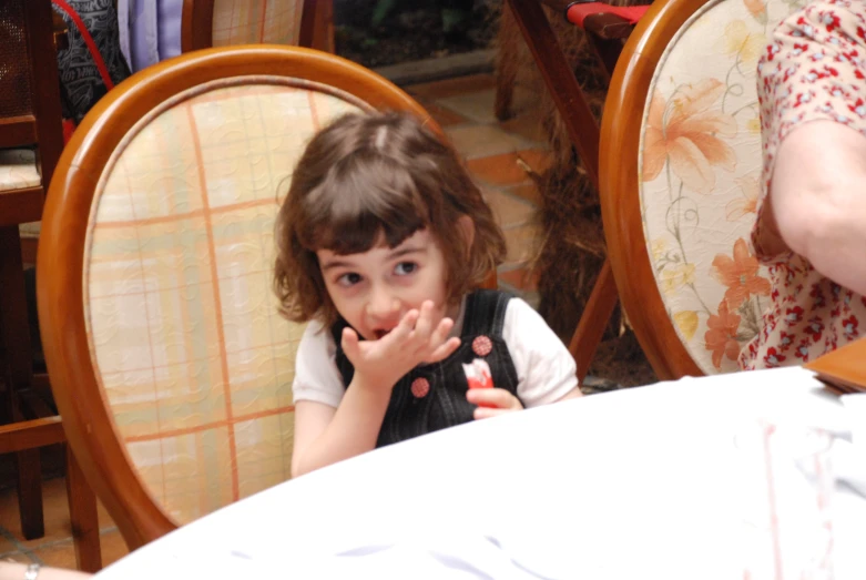 a little girl sitting in a chair holding her hand up to her mouth