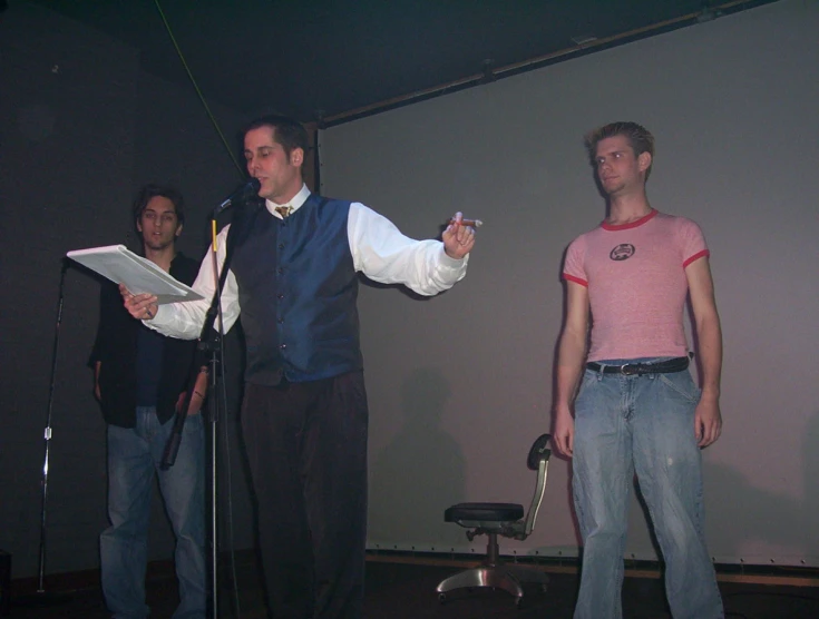 three men stand in front of a microphone while one man holds up a piece of paper