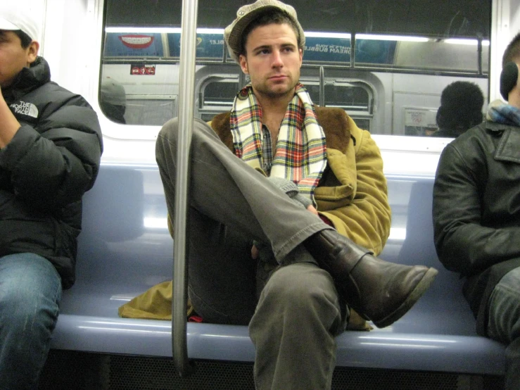 man sitting on bus with blanket covering his head and one foot near mouth