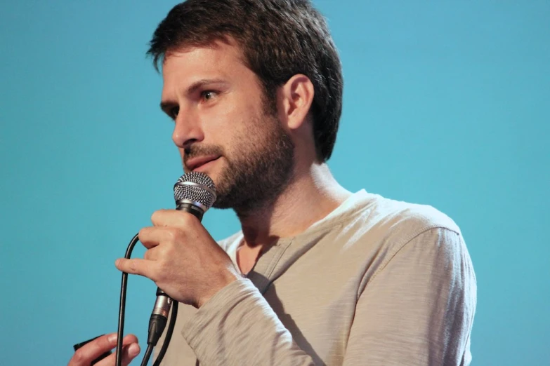 a man with short hair wearing a beige shirt holds a microphone and speaks