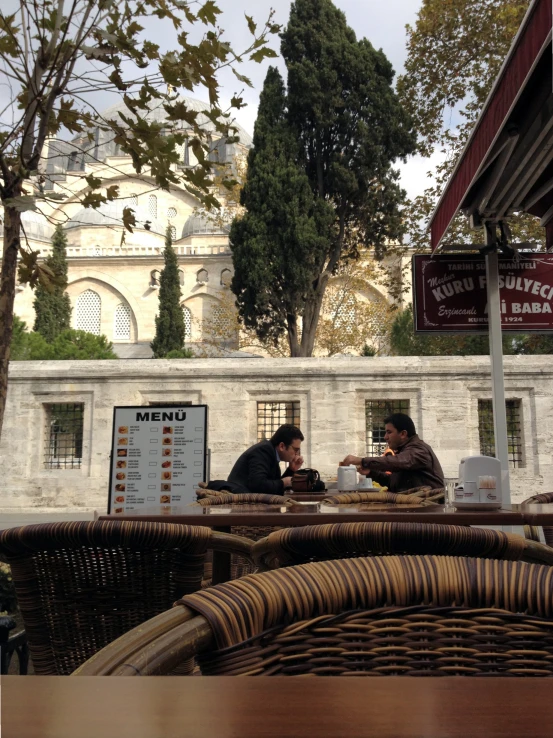 two men sitting at tables outside next to some trees